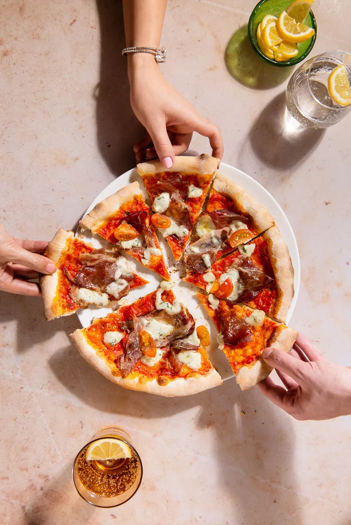 Hands reaching for slices of pizza, with drinks and plates on a sunlit table.