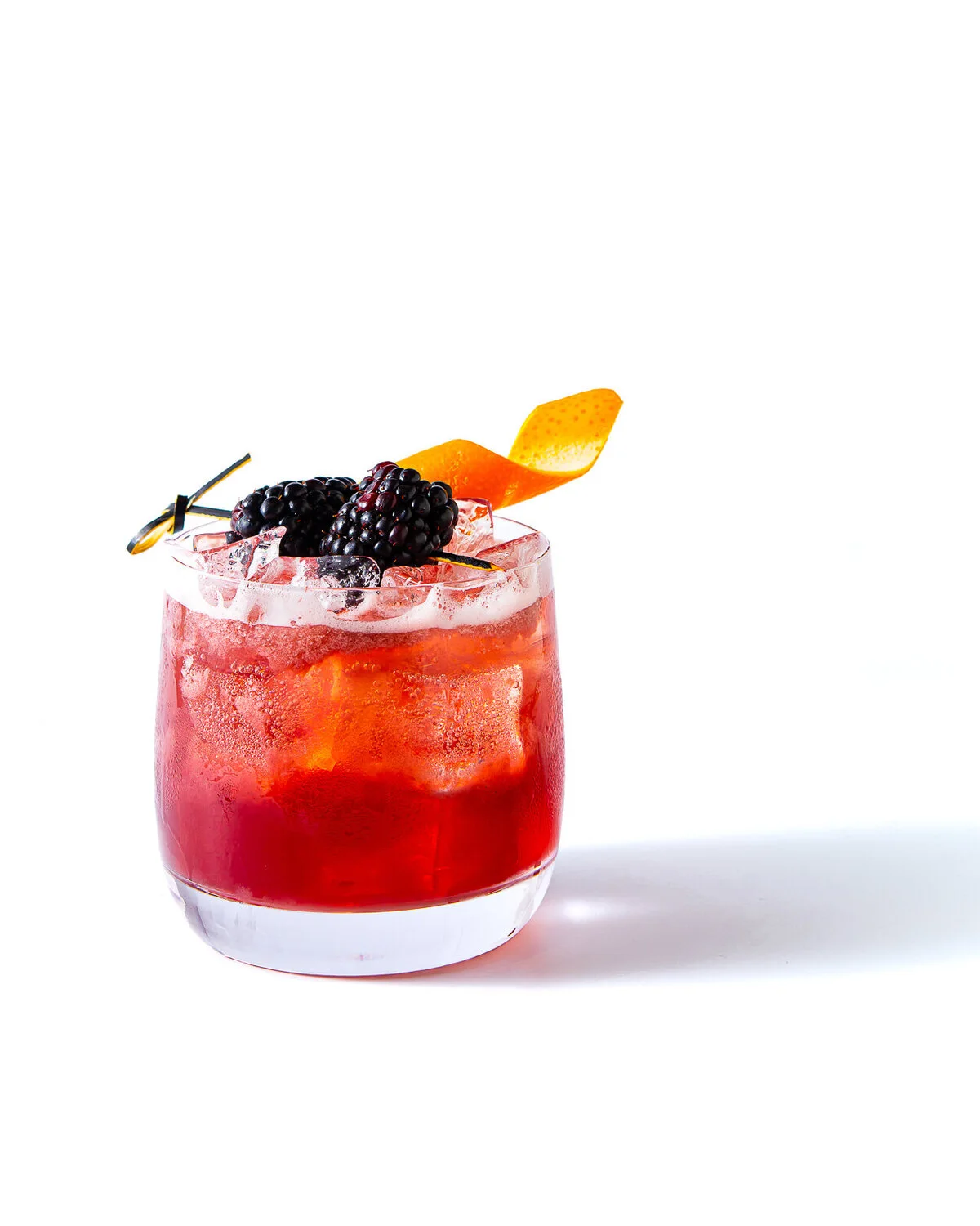 Red cocktail with berries and a citrus garnish against a white backdrop.