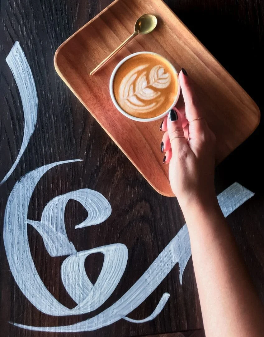 Hand holding coffee on a tray over a table with Arabic calligraphy.