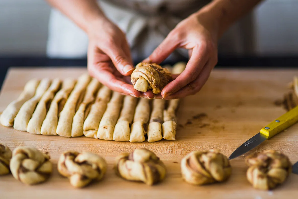 Hands shaping a cinnamon bun above rows of prepared dough.