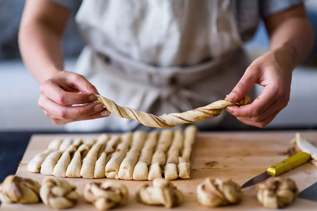Hands twisting dough above rows of prepared cinnamon buns.