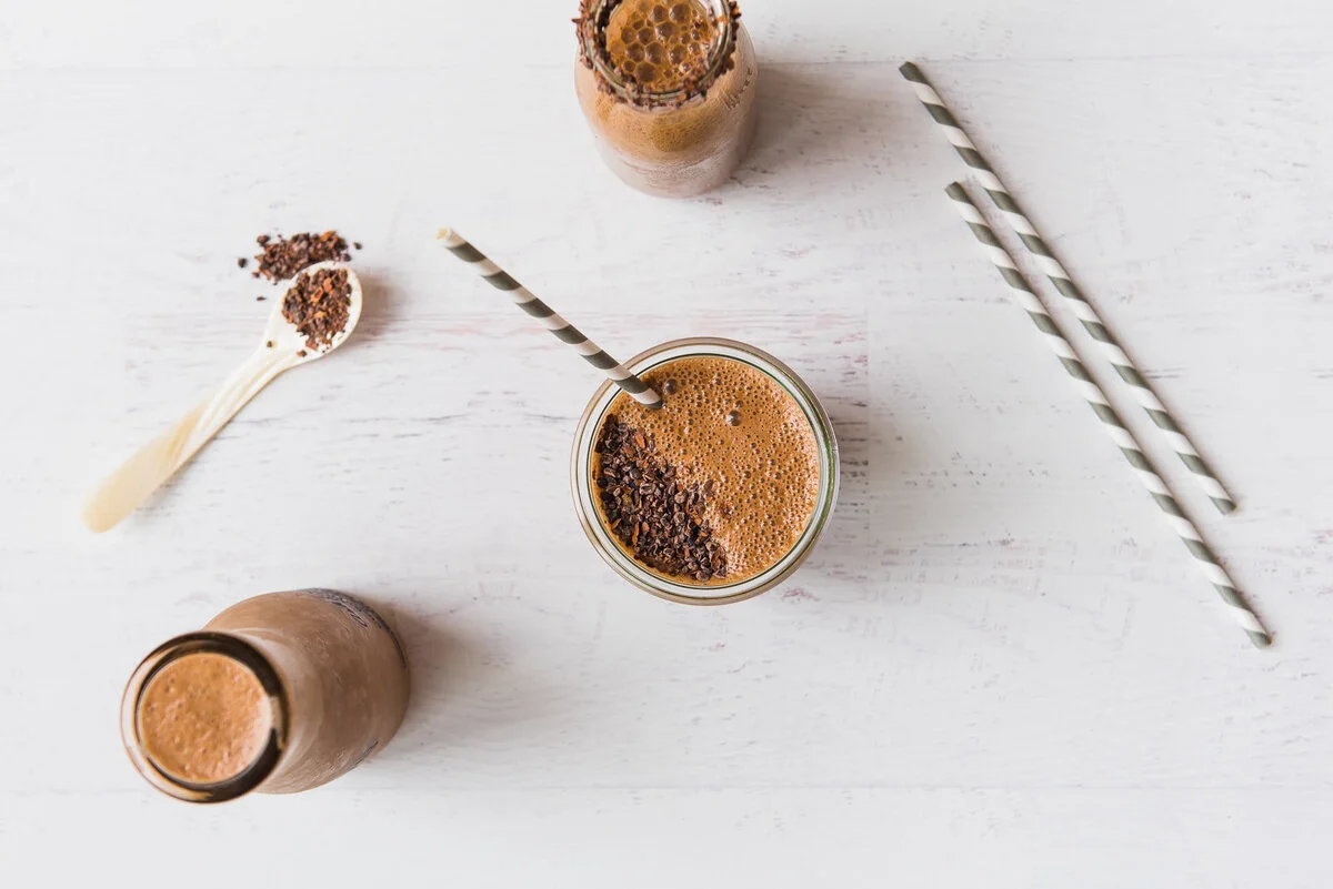 Glasses of cacao almond milk with chocolate nibs on a pale tabletop.