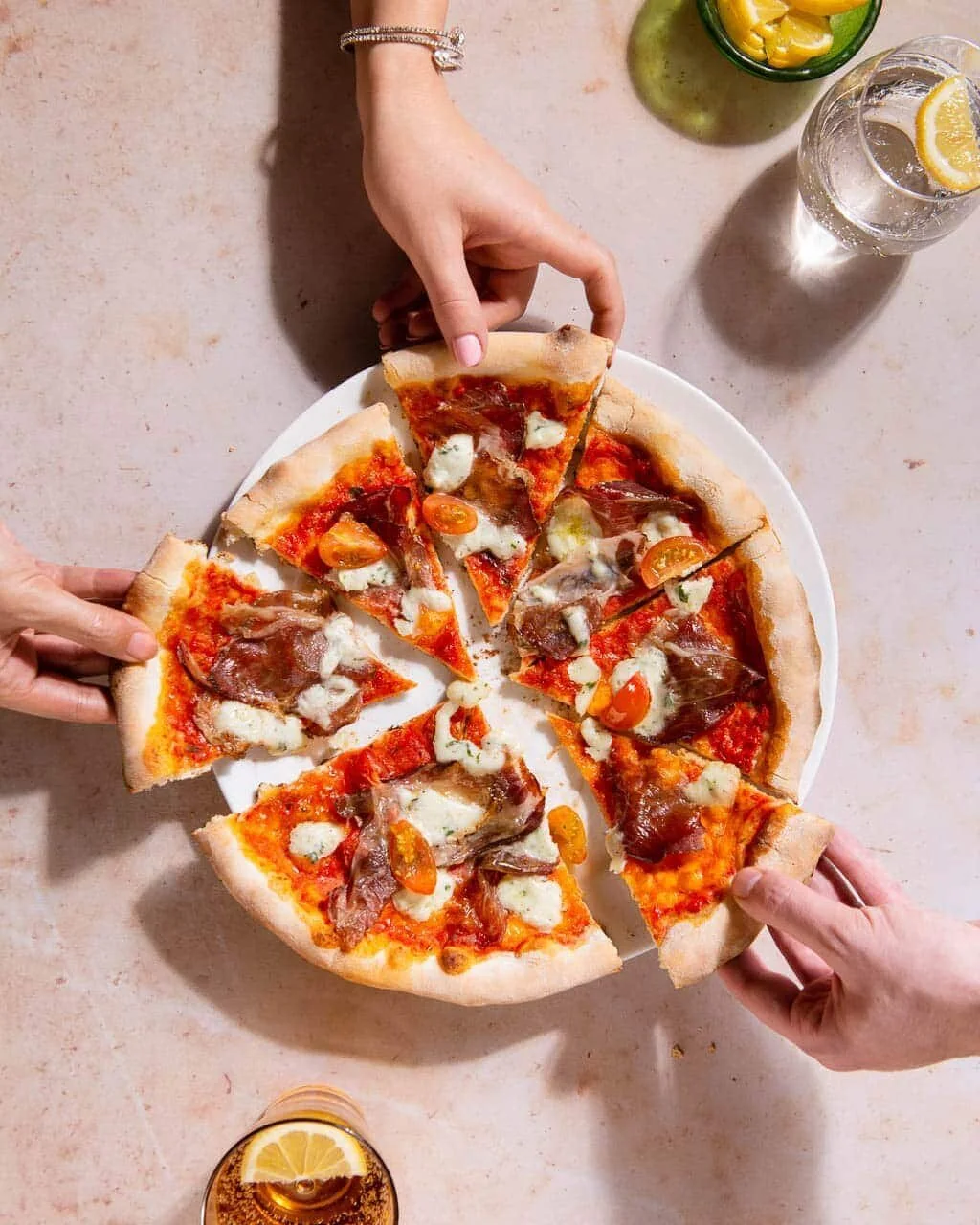Hands reaching for a pizza on a pale tabletop.